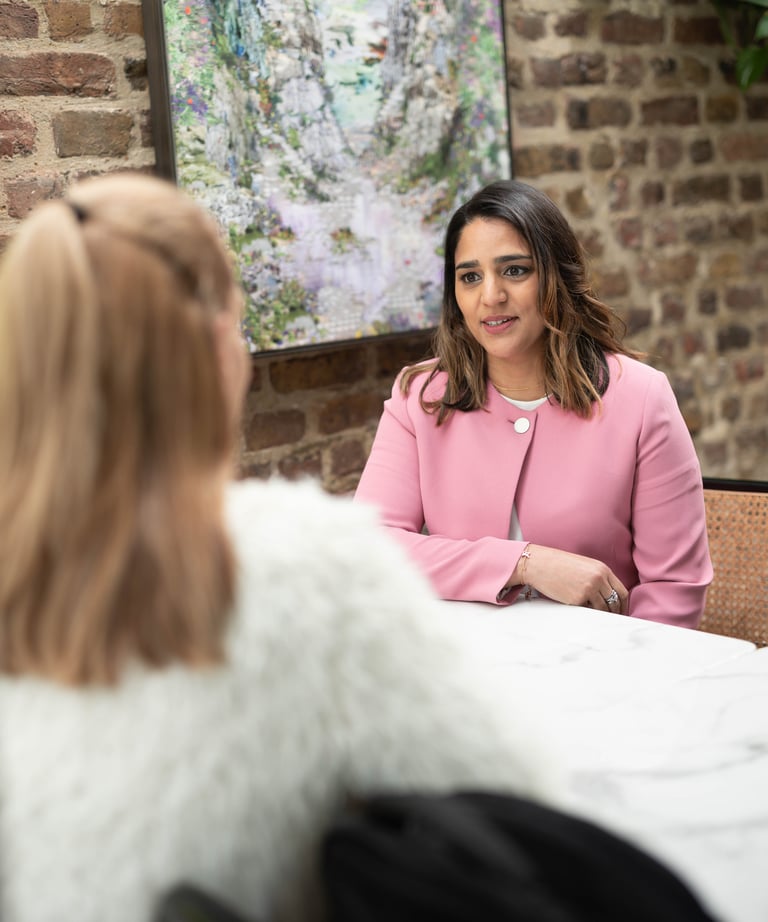 A photo of Anita wearing a pink jacket sitting at a table talking to a person who has their back to the camera.