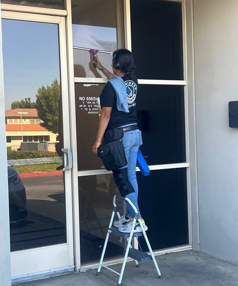 Professional window cleaner using a squeegee on a commercial glass door while standing on a ladder.