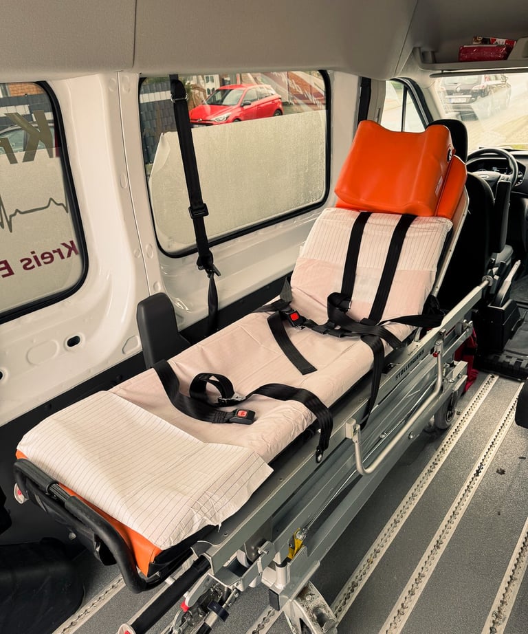 Trage im Rettungswagen mit orange Kopfstütze, rotes Auto draußen.