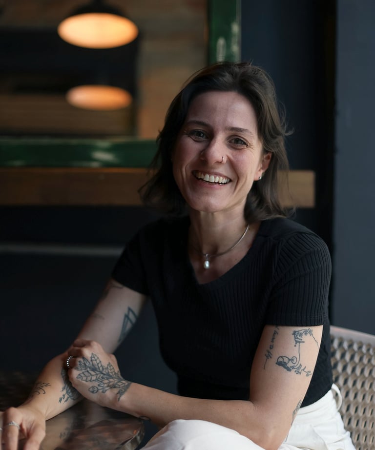 Smiling woman with tattoos sitting at a cafe table, wearing a black shirt and white trousers.