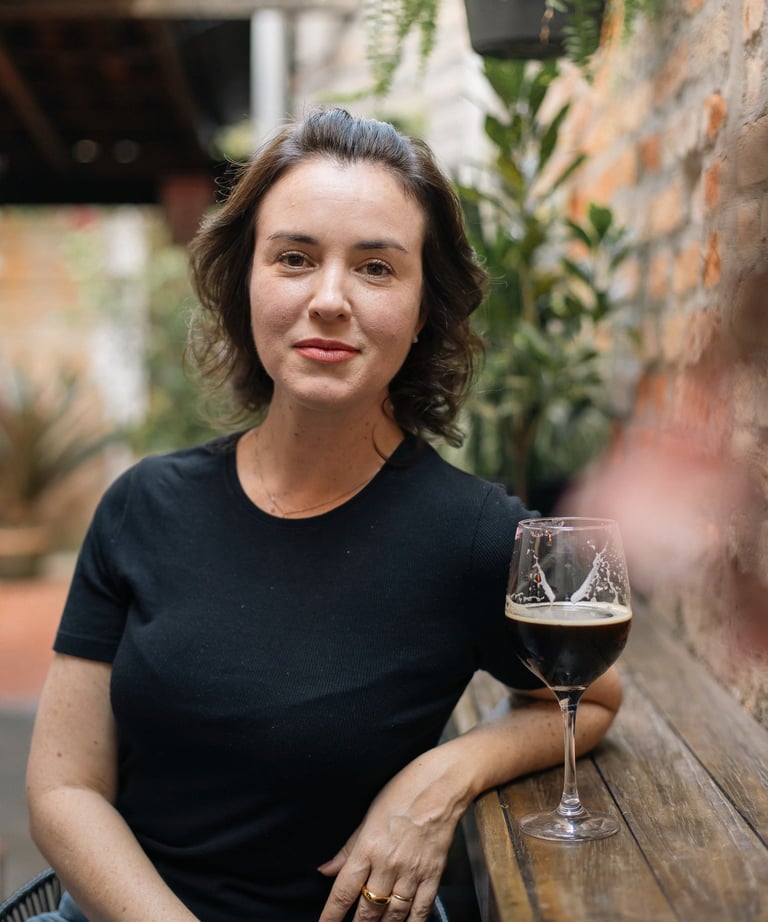A woman in a black t-shirt sitting at a wooden patio table with a glass of dark beer.