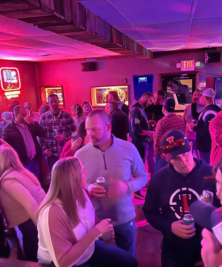 Crowded local bar with patrons enjoying drinks under red and blue neon beer signs.
