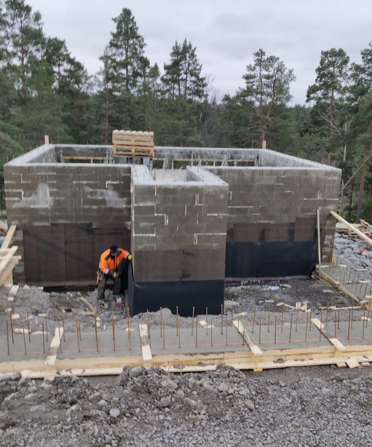 A construction worker builds a concrete block foundation for a new house in a forest setting.