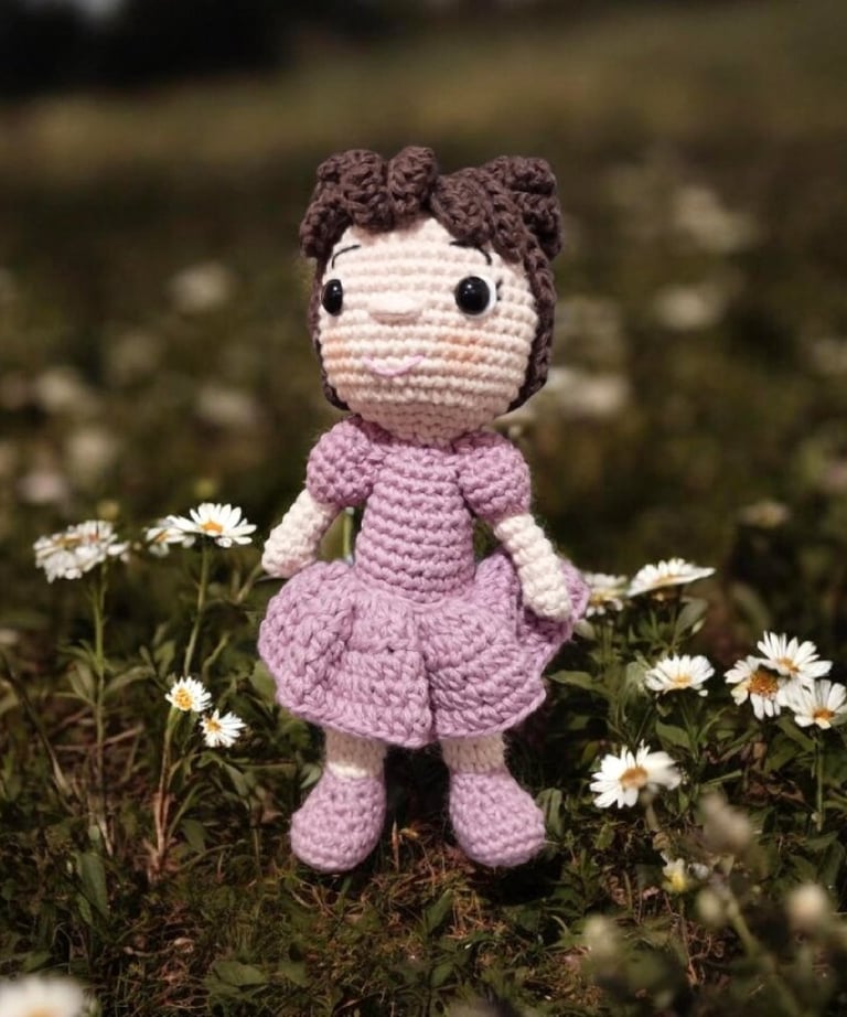 Muñeca amigurumi de ganchillo hecha a mano, vestido morado, de pie en un campo de margaritas blancas