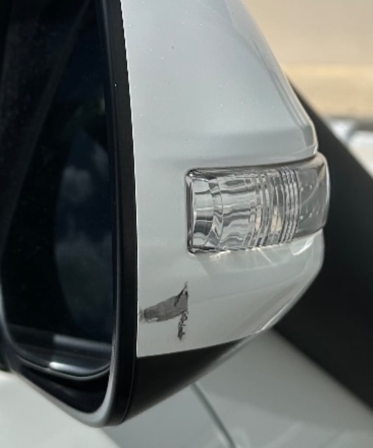 Close-up of a white car side mirror with a scratch and paint damage near the turn signal light.