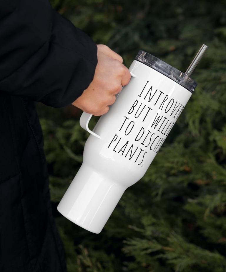 Hand holding white plant-themed travel mug outdoors near green foliage.
