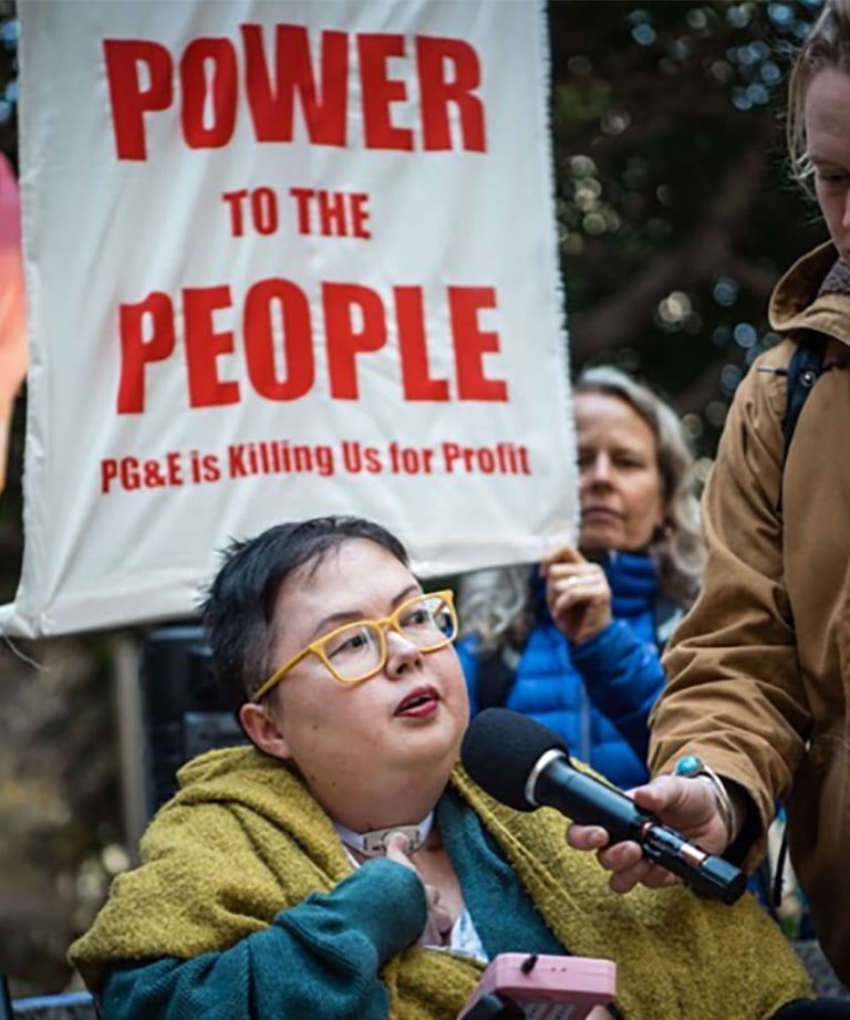 Photo of Stacey at a protest with sign that reads "power to the people PG&E is killing us for profit"