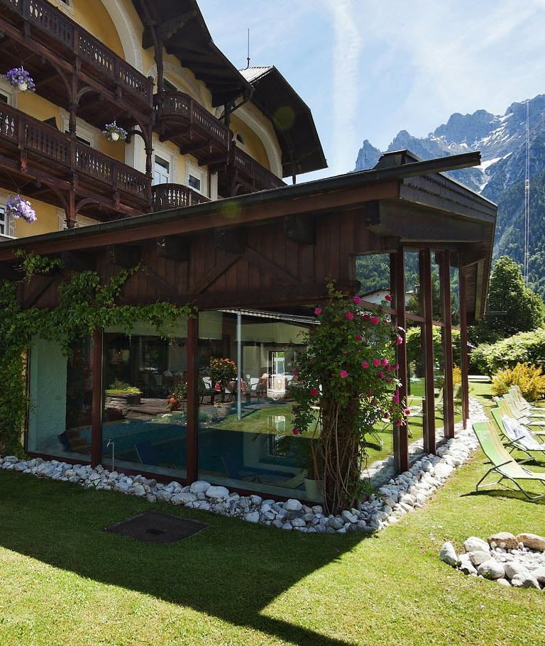 Luxury alpine hotel with an indoor pool and sun loungers on a green lawn facing the Alps.