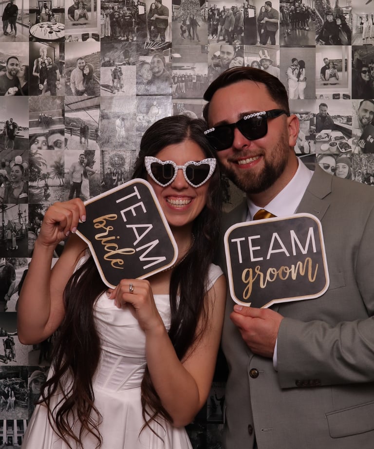 Bride and groom posing in a photo booth with fun signs and heart-shaped sunglasses.