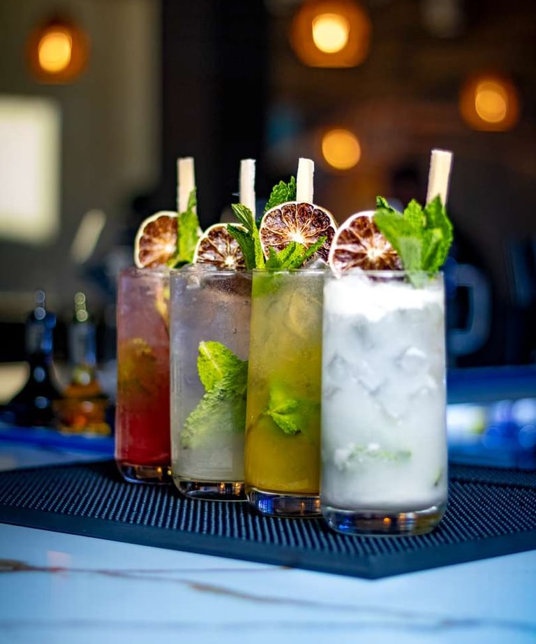 Variety of colorful mojito cocktails with fresh mint and dried lime garnishes on a bar mat.