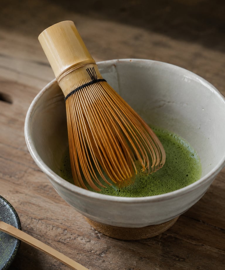 Traditional bamboo whisk stirring frothy green matcha tea in a ceramic bowl on a wooden surface.