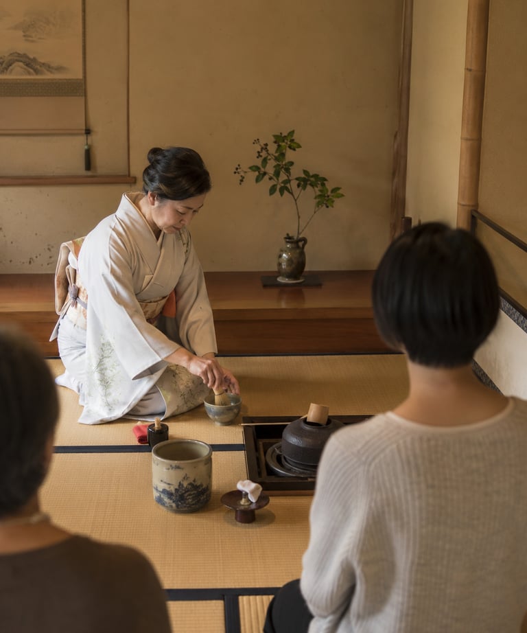 A Japanese woman in a kimono performs a traditional matcha tea ceremony for guests on tatami mats.
