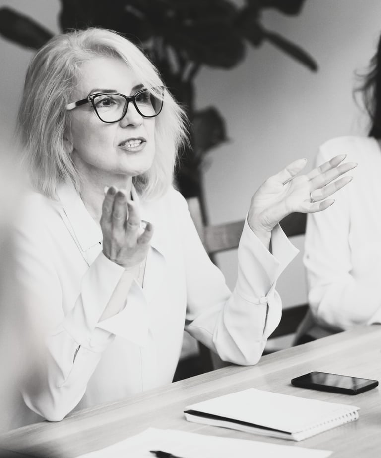 a woman in a white shirt and glasses sitting at a meeting table gesturing with her hands