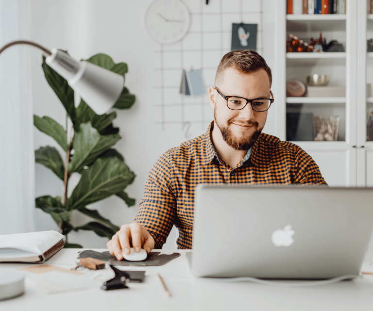 Smiling man with glasses working on a laptop at a bright home office desk.