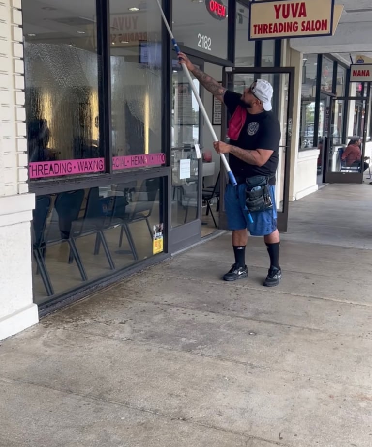 Professional window cleaner using a long extension pole to wash the glass storefront of a local business.