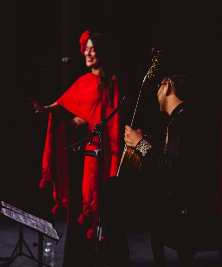 A Spanish flamenco dancer in a red shawl performs on stage with a live acoustic guitarist.