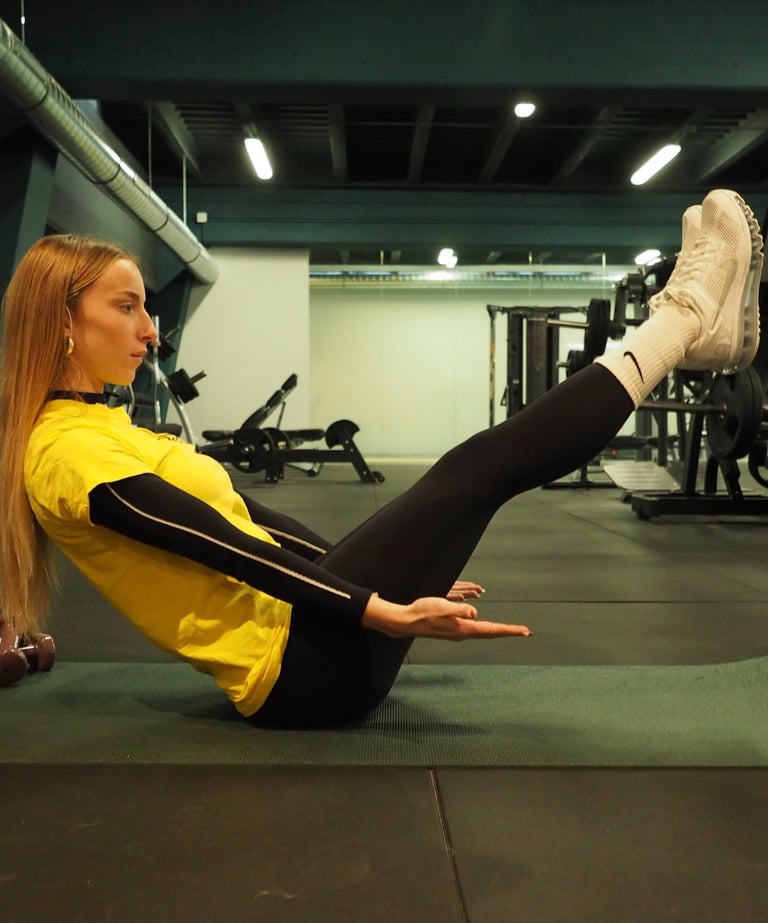 Una donna in maglia gialla esegue l'esercizio 'boat pose' su un tappetino da yoga in palestra, concentrandosi sull'equilibrio