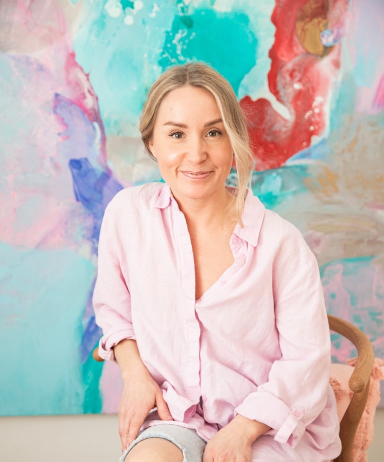 Saija Uussaari in a pink shirt and jeans sitting on a chair in front of a colorful painting