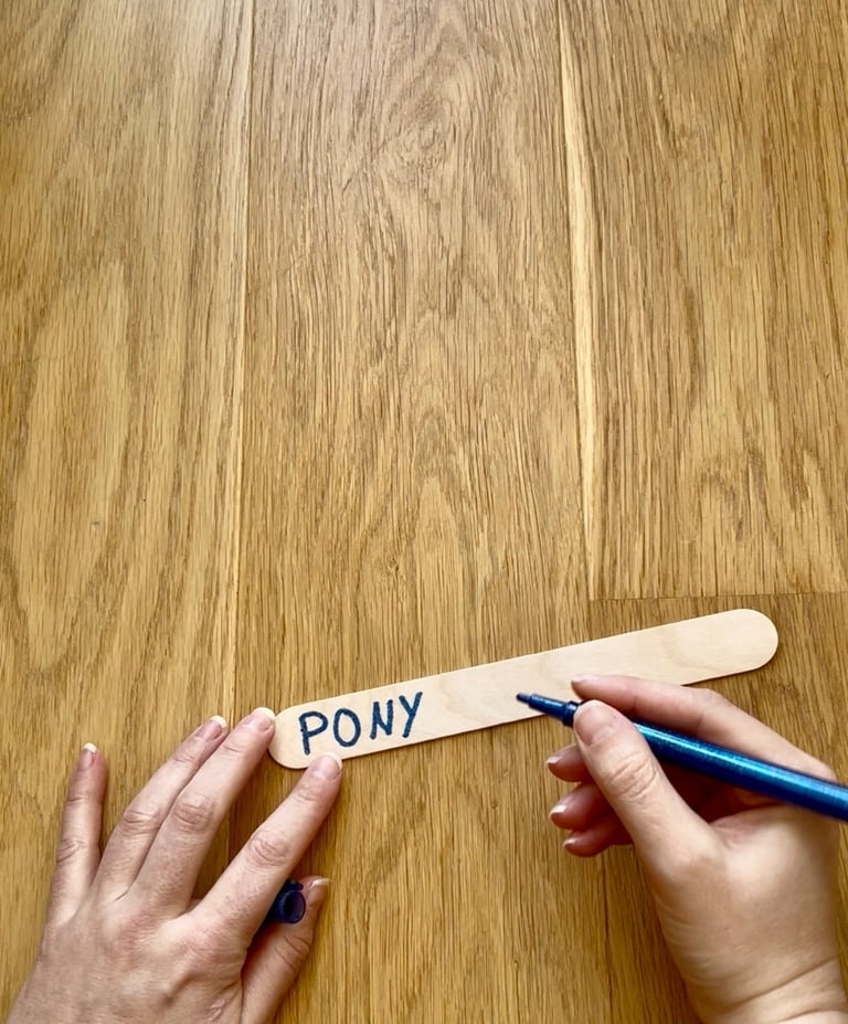 Hand writing the word PONY with a blue marker on a wooden popsicle craft stick.
