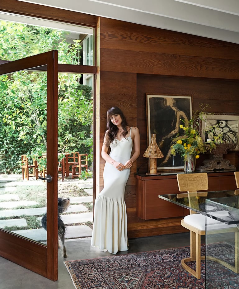 Dakota Johnson in white gown posing by glass doors at home.