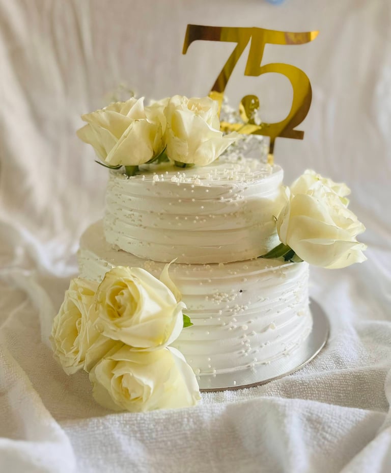 Two-tier white buttercream 75th birthday cake decorated with fresh white roses and a gold topper.