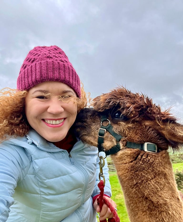 Smiling woman in a purple beanie taking a selfie with a fluffy brown alpaca on a farm in Ireland.