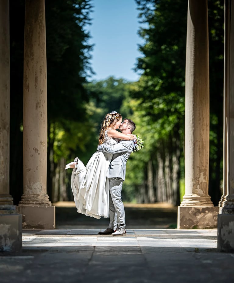 Bräutigam hebt seine Braut lachend in die Höhe – romantischer Hochzeitsmoment in Mailand unter alten Säulen.