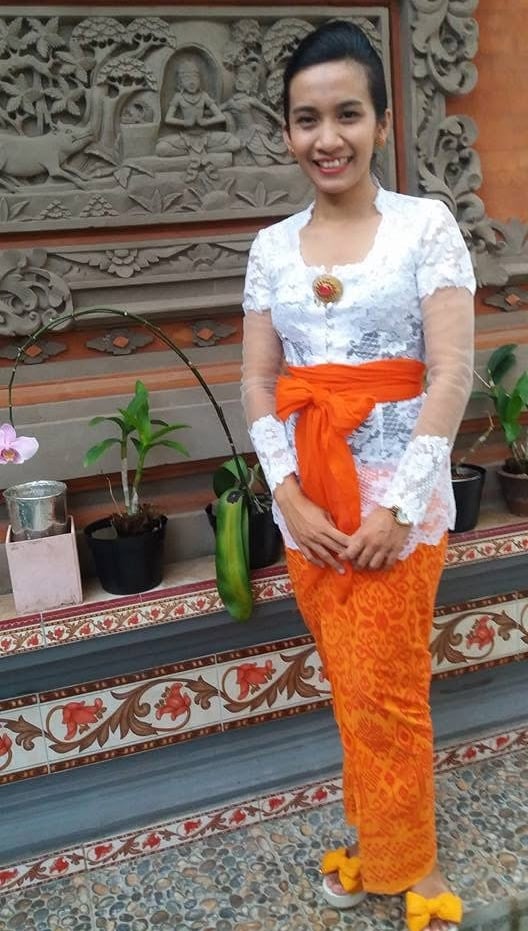 A Balinese woman wearing a white lace kebaya and orange sarong stands before a carved stone wall.