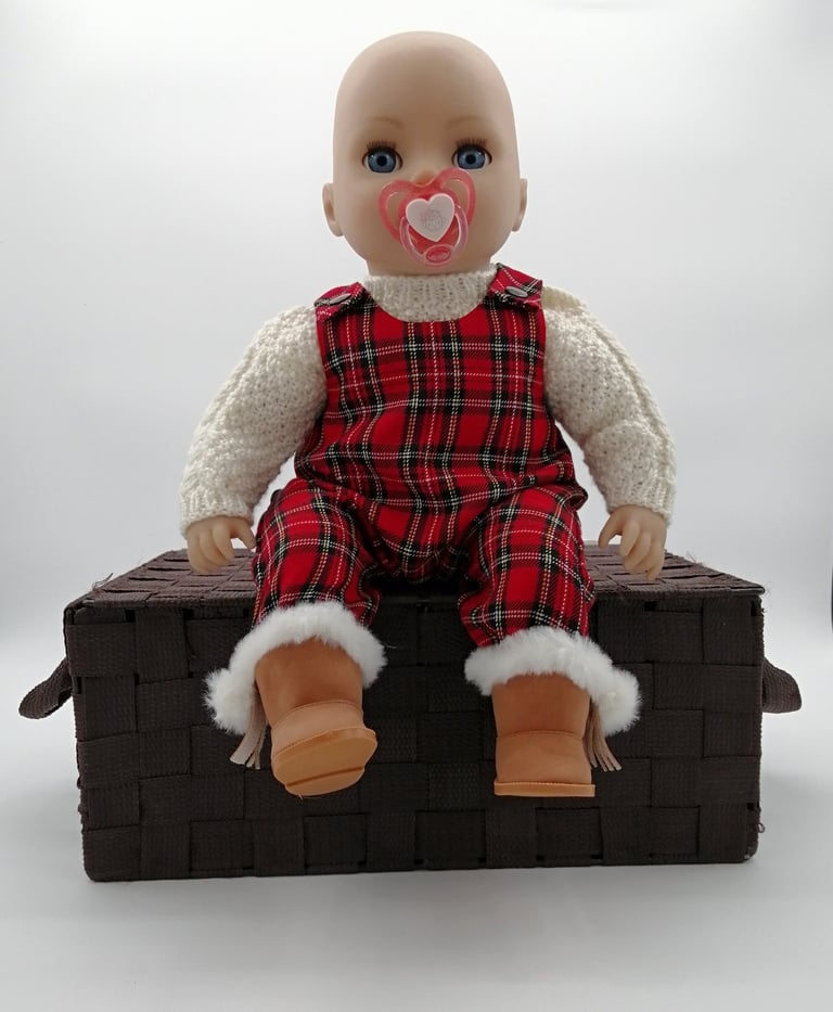 a Baby Annabell baby doll sitting on a basket dressed in handmade tartan overalls