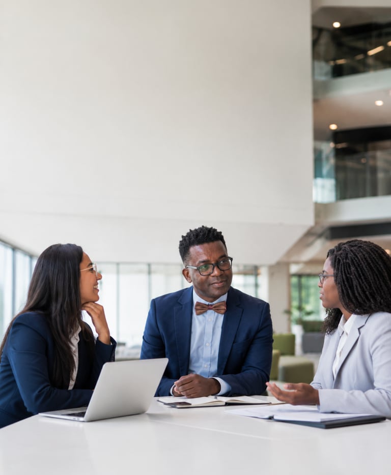 Three diverse business professionals in formal attire having a meeting at a white office table with a laptop.