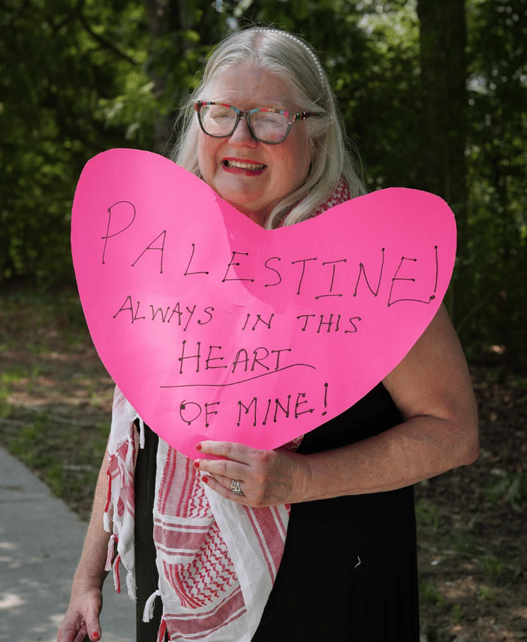 a woman wearing a keffiyeh stands holding a sign that says "palestine! always in this heart of mine"