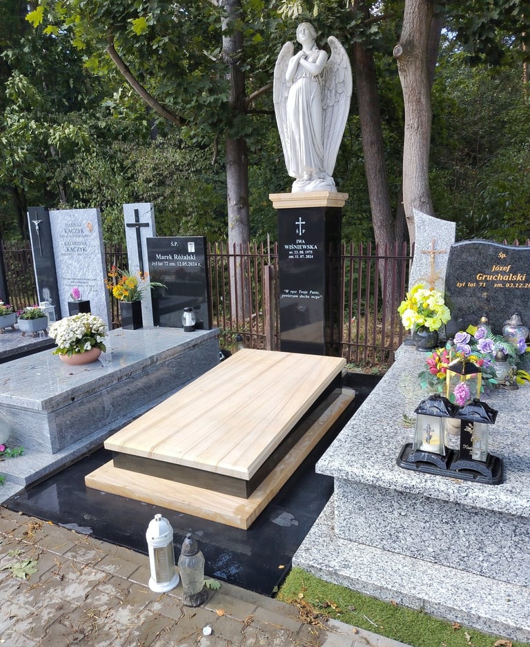 Polished granite tombstones and a white angel statue in a cemetery with floral tributes.