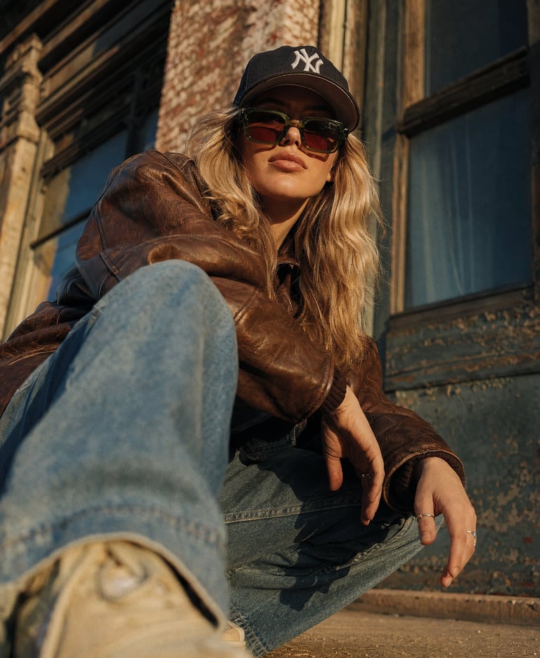 A blonde woman in a brown leather jacket, sunglasses, and baseball cap posing in urban street style.