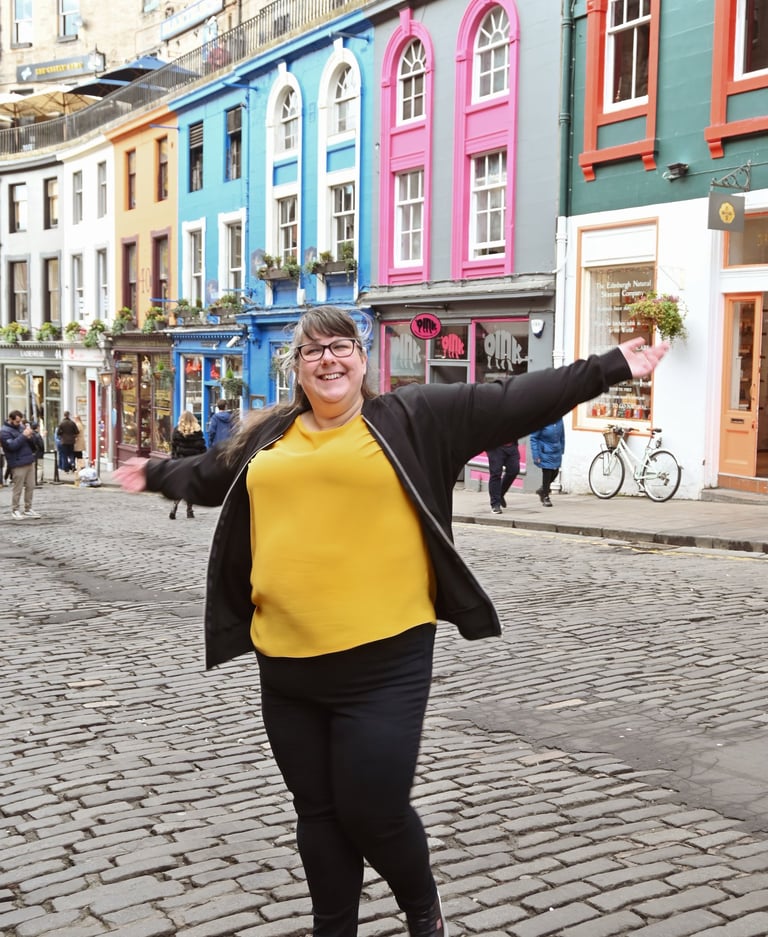 Laura is dancing with her arms out on a cobbled street with brightly coloured buildings behind her