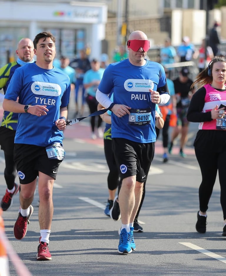Two runners side by side, one is blindfolded, both are holding a tether for a charity fundraising ru