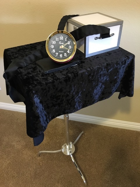 Magician table with a black velvet cloth, analog clock, and mystery box prop for stage magic.