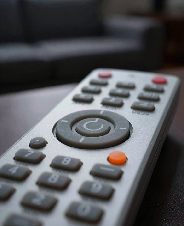 Crisp macro photography of a physical TV remote control, focused on the power button and orange indicator LED, moody living room ambient light, shallow depth of field, 50mm.