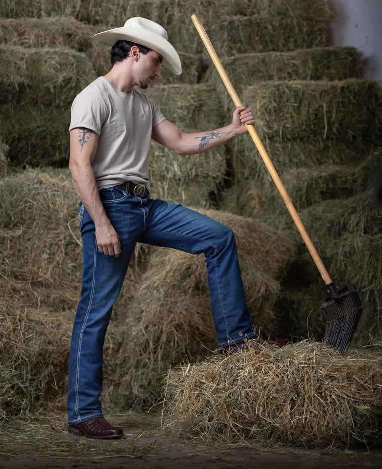 A ranch hand in a white cowboy hat and blue jeans holding a pitchfork in front of hay bales.