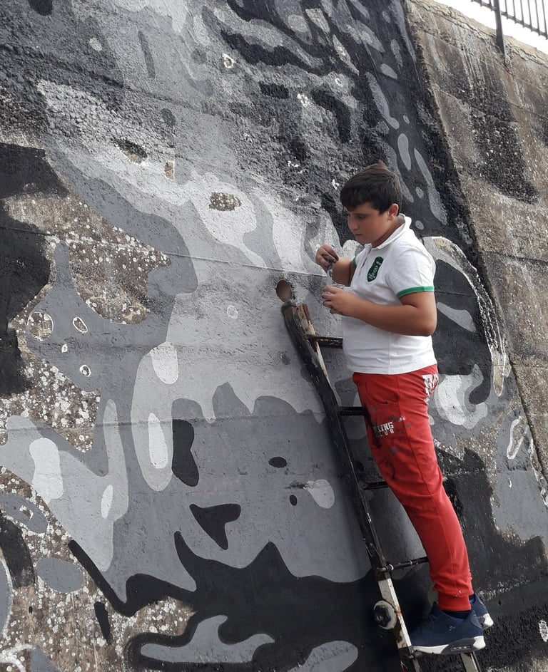 Young artist painting a grayscale mural on an outdoor wall, project curated by Poliops.