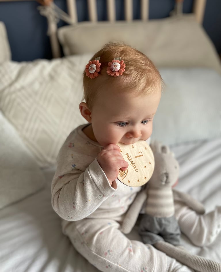 Niemowlę siedzi na łóżku, na głowei ma spinki do włosów w kształcie kwiatów