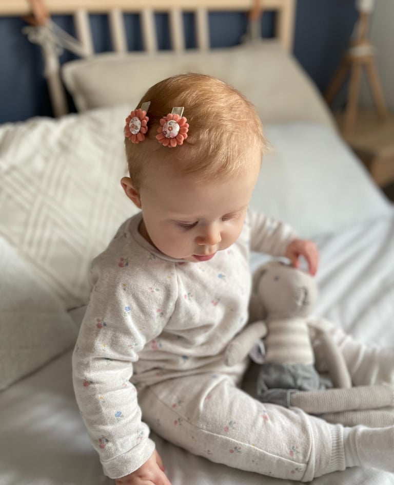 niemowlę siedzi na łóżku z pluszową zabawką, na głowie ma dwie spinki w kształcie kwiatów