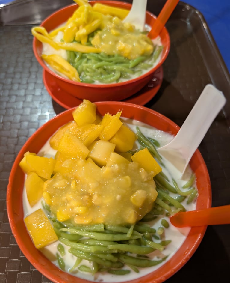 Two bowls of Malaysian Cendol dessert with green pandan noodle, fresh mango, corn, and coconut milk.