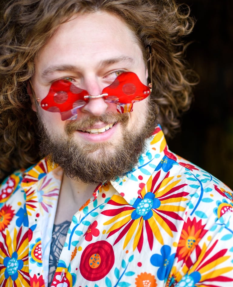 A smiling man with a beard wearing mushroom-shaped red sunglasses and a colorful floral shirt