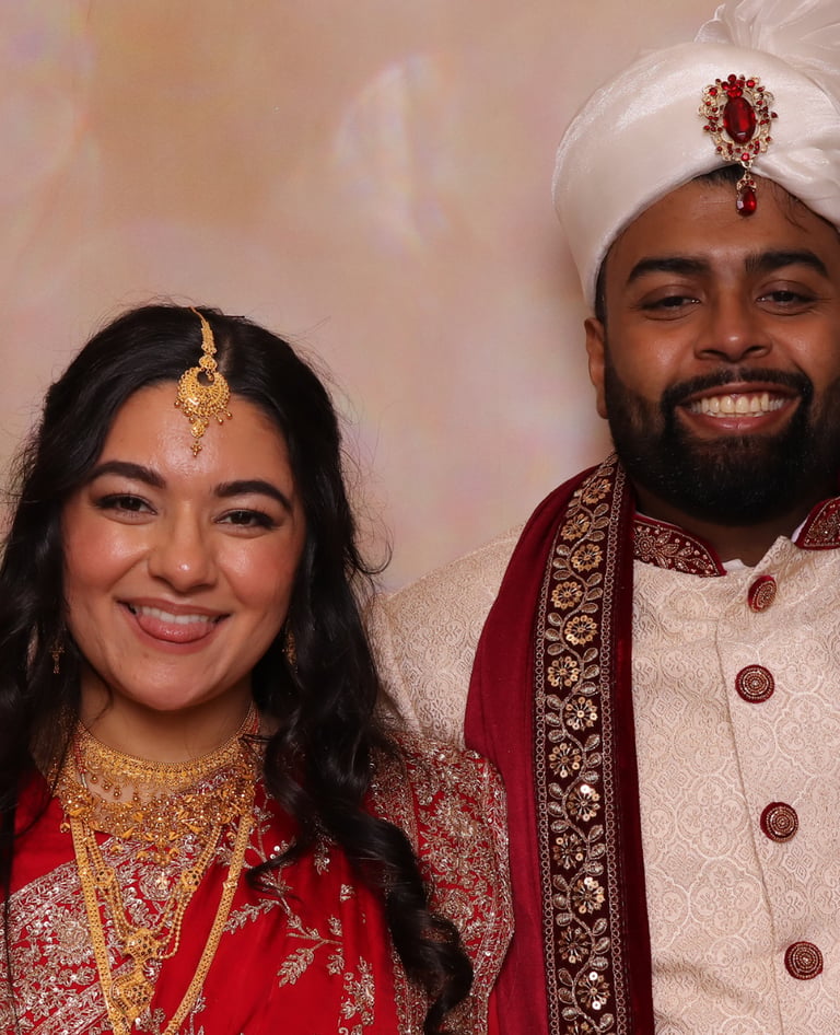 A smiling South Asian couple in traditional wedding attire, featuring a red gold-embroidered sari and a white sherwani
