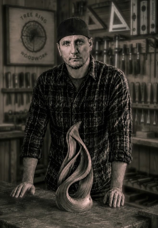Owner of Tree Ring Woodwork, Isaac Griffin, in front of a wooden sculpture in a workshop.