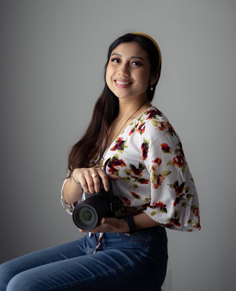Professional shot of me holding Canon camera, sitting on a stool. Wearing floral top and headband.