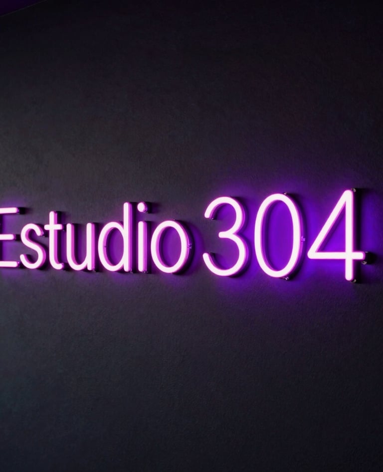 A sleek dark wall featuring the name 'Estudio 304' glowing in vibrant neon pink and neon violet lights, premium modern adult entertainment studio design, dark-mode styling.