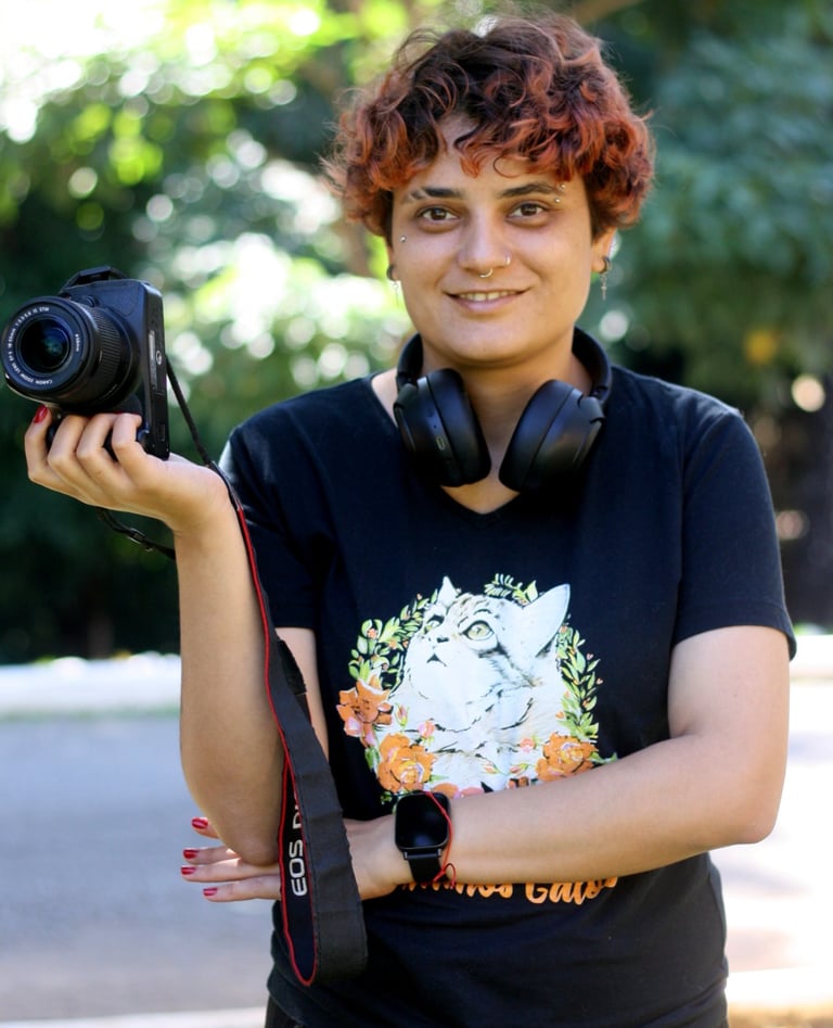 A smiling young female photographer with short curly red hair holds a DSLR camera, wearing headphones around her neck and a
