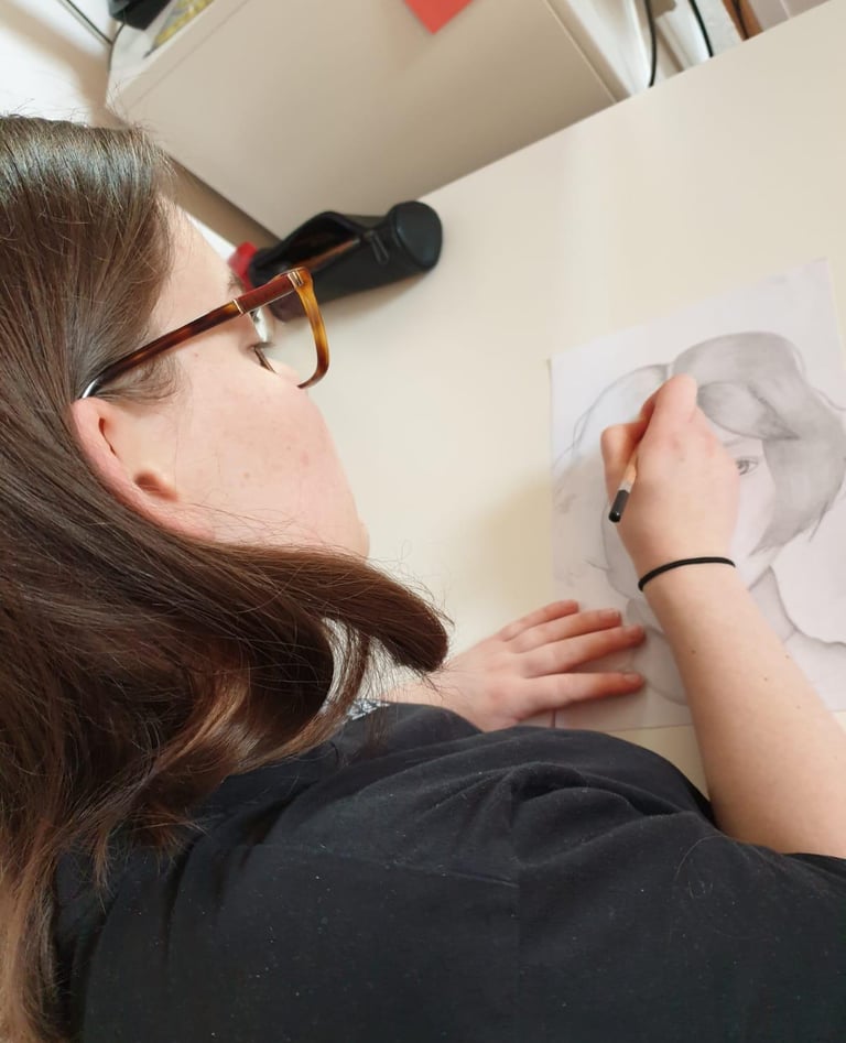 A woman with glasses focused on drawing at a desk, surrounded by art supplies.