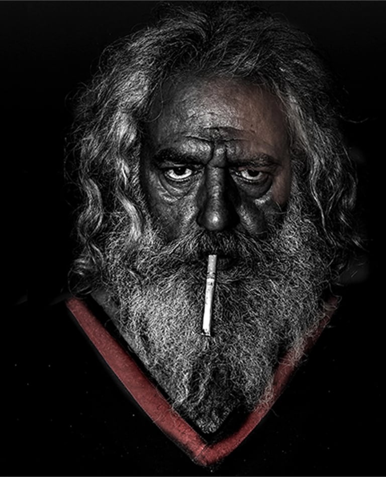 Portrait of an older man with a weathered face and intense gaze, cigarette held between his lips. 